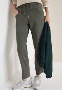 Personne portant un pantalon à cordon de serrage vert olive et des baskets blanches, tenant une veste vert foncé dans la main droite.