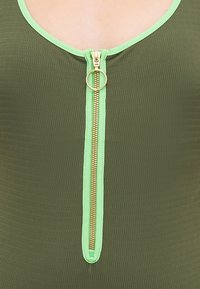 Maillot de bain vert olive avec une texture côtelée, une fermeture éclair dorée à l'avant avec un tirette circulaire, et un passepoil vert clair contrastant le long de l'encolure.