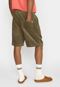 Olijfgroene corduroy shorts met een elastische tailleband, zijzakken en een gestructureerd ribbelpatroon. Geregisseerd met witte sokken en bruine sandalen.