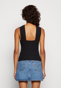 Mujer con cabello rizado que lleva una blusa sin mangas negra con la espalda baja y una falda de mezclilla azul, de pie frente a un fondo claro y sencillo.