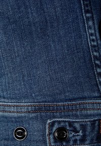 Mörkblå denimtyg med en subtil textur, med orange sömmar och två svarta metallknappar nära botten.