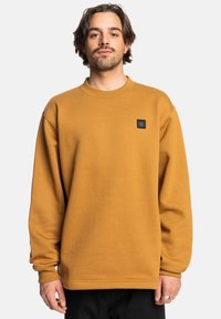 Sudadera color mostaza hecha de tela suave, con cuello redondo y un pequeño parche de logo negro en el pecho. Corte holgado con mangas largas.