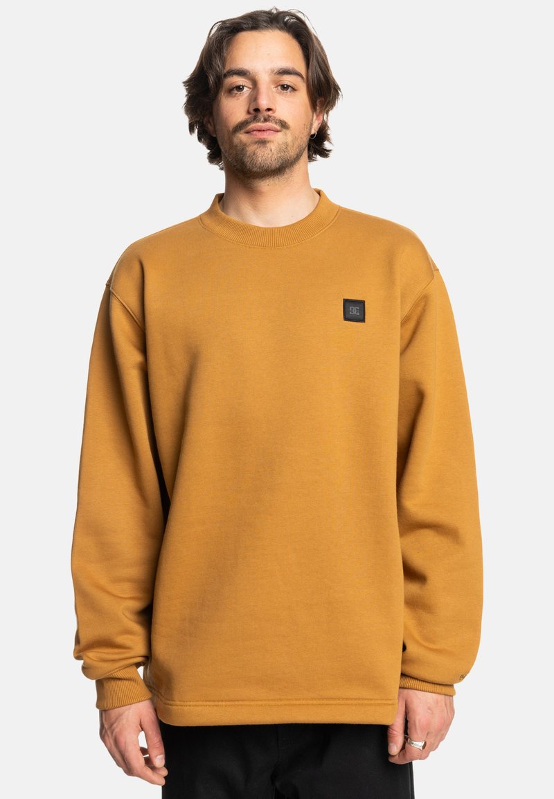 Sudadera color mostaza hecha de tela suave, con cuello redondo y un pequeño parche de logo negro en el pecho. Corte holgado con mangas largas.