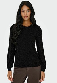 Svart langermet topp med teksturert bølgemønster, rund hals og tett i ermene, kombinert med brune bukser.