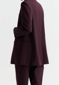 Vínový oblek s přizpůsobeným sakem s výrazným klopovým límcem, zapínáním na jeden knoflík a kalhotami s přímým střihem; hladká, texturovaná látka.