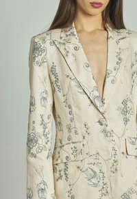 Blazer in lino color crema con ricami floreali blu e verdi, caratterizzato da un profondo scollo a V, maniche lunghe, due tasche frontali e chiusura con bottone.