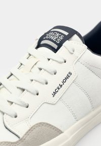 Witte sneaker met grijze suède accenten, bovenwerk van canvas en leer, ronde neus, platte veters en merklabel op de tong. Gestructureerde zijpanelen.
