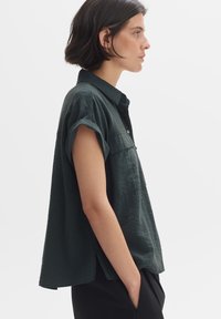 Camicia a maniche corte, di colore verde scuro, con una vestibilità morbida, colletto, due tasche frontali e tessuto testurizzato. Fessure laterali all'orlo.