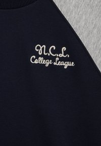 Tamsiai mėlynas džemperis su pilka peties juosta, siuvinėta baltais rašmenimis „N.C.L. College League“ stilizuotu raštu. Klasikinio dizaino apvalus kaklas.