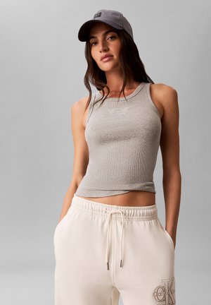 Kobieta w szarym topie, jasnoszarej czapce i kremowych spodniach dresowych Calvin Klein z rękami w kieszeniach, stojąca na jednolitym tle.