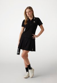 Schwarzes Poloshirt mit weißen Streifen, Logo auf der Brust, kombiniert mit einem schwarzen Rock. Das Model hält eine schwarze Wasserflasche. Weiße Sneaker und schwarze Socken.