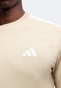 Μπεζ πουκάμισο με λευκό λογότυπο Adidas στο αριστερό στήθος και λευκές ρίγες στον ώμο, φορεμένο από ένα άτομο με σκουρόχρωμο δέρμα.