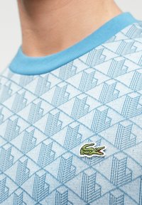 Ανοιχτό μπλε υφασμάτινο πουκάμισο με γεωμετρικό σχέδιο και αντίθεση μπλε γιακά. Διαθέτει ένα μικρό πράσινο λογότυπο κροκόδειλου στο μπροστινό μέρος.