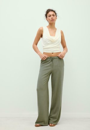 Giovane donna in pantaloni larghi verde oliva e top crema senza maniche drappeggiato, in piedi davanti a uno sfondo chiaro e uniforme.