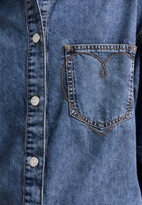 Denim krekls gaiši zilos toņos, ar sudraba pogām un sirds formas kabatu ar kontrastējošu oranžu diegu šuvēm.