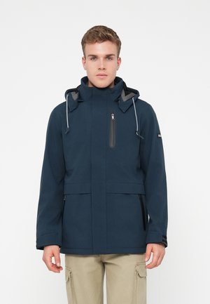 Jovem vestido com uma jaqueta com capuz azul-marinho e bolsos com fecho, e calças cargo bege, de pé contra um fundo branco.