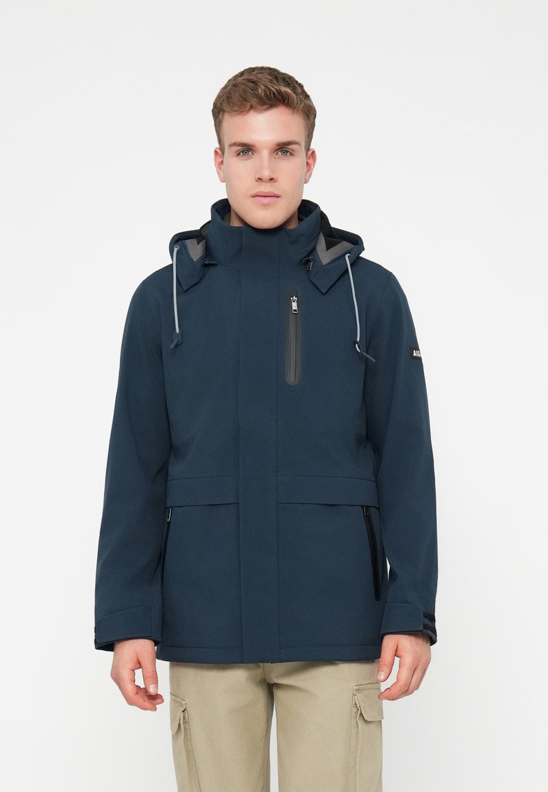 Νεαρός άνδρας φορέσει μια ναυτική μπλε μπουφάν με κουκούλα και φερμουάρ τσέπες, και μπεζ cargo παντελόνι, που στέκεται μπροστά σε άσπρο φόντο.