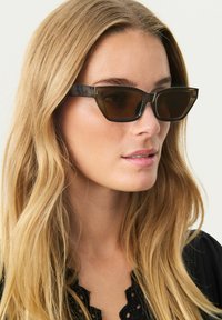 Lunettes de soleil teintées marron avec une monture noire géométrique et une forme angulaire, présentant une texture brillante et de discrets accents matériels.