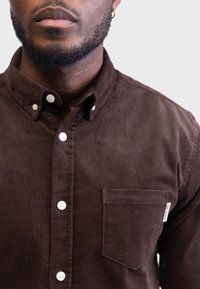 Brązowa koszula z korduroju z kołnierzykiem zapinanym na guziki, białymi guzikami oraz jedną kieszenią na piersi z metką marki. Gładka tekstura i dopasowany krój.