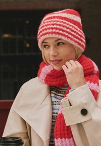 Red and pink striped knit beanie and matching scarf, soft texture, layered over a patterned sweater and a beige coat with black buttons.