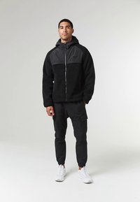 Chaqueta de forro polar negra con un panel gris, cremallera y capucha, combinada con pantalones cargo negros y zapatillas blancas. Material texturizado y ajuste casual.