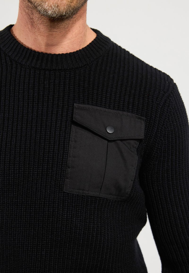 Maglione nero a maglia con un motivo testurizzato, dotato di una tasca sul petto in tessuto nero con bottone a pressione. Collo rotondo, polsini a costine.