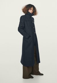 Cappotto di lana blu navy con colletto alto, maniche lunghe e chiusure a toggle. Presenta tasche laterali e una silhouette dritta.