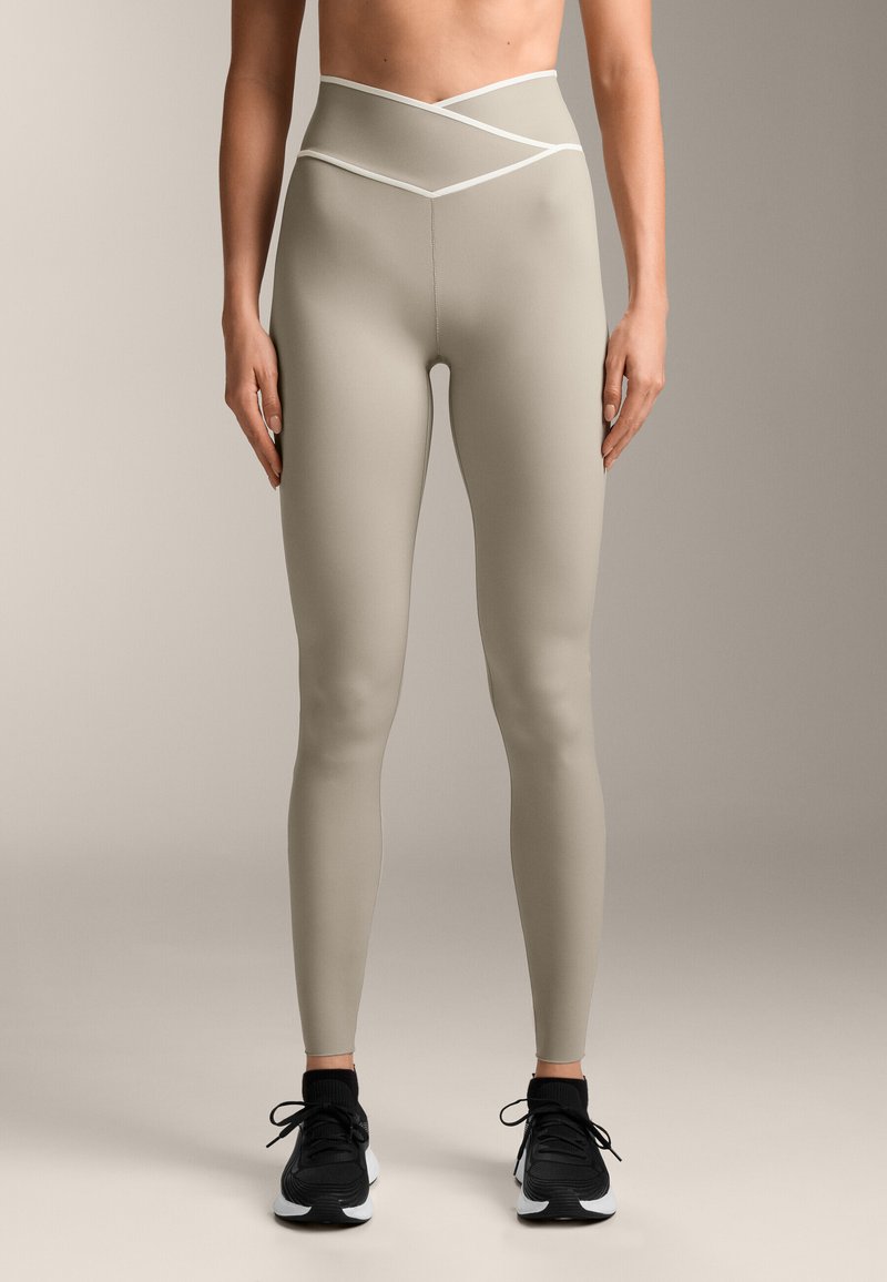 Magasan fazonú, világos bézs leggings, keresztezett derékpánttal, rugalmas anyagból, sima textúrával, fekete sportcipővel párosítva.