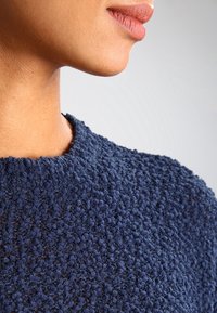 Gros plan sur le bas du visage et le cou d'une personne portant un pull en maille texturée bleu marine, sur un fond clair uni.