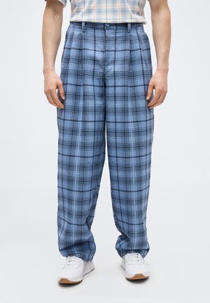 Homme portant un pantalon ample à carreaux bleus, associé à des baskets blanches et une chemise à carreaux rentrée, debout devant un fond uni.