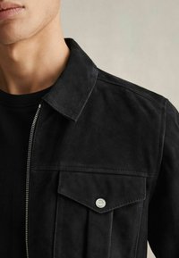Veste en daim noir avec col, fermeture éclair sur le devant et une poche poitrine dotée de garnitures métalliques. Surface texturée avec peu de détails.
