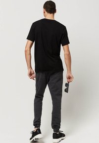 Sort bomulds t-shirt, grå joggingbukser og sorte sneakers. Holder solbriller. Set fra ryggen med enkelt design og afslappet pasform.