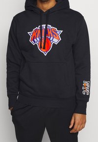 Sweat à capuche noir avec logo New York Knicks à l'avant, présentant des accents orange et bleu. Comprend une poche kangourou et le texte NYC sur la manche.