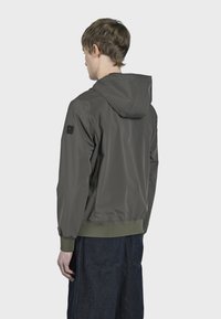 Tamno maslinasto zeleni dukserica s kapuljačom i elastičnim manžetama i rubom. Ima oznaku na lijevom ramenu i glatku, sjajnu teksturu.