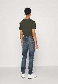 Schmal geschnittene dunkelblaue Jeans mit einer verblassten Textur, kombiniert mit einem grünen T-Shirt mit kurzen Ärmeln und weißen Sneakers, von hinten betrachtet.