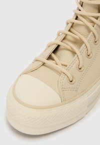 Detailní záběr na béžovou koženou tenisku s odpovídajícími tkaničkami a texturovanou gumovou podrážkou na bílém pozadí.