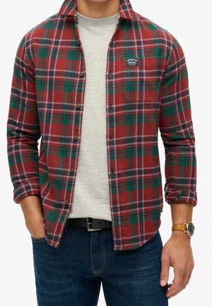 Mann trägt ein rot-grün kariertes Hemd über einem hellgrauen Pullover, dunkelblaue Jeans und eine braune Armbanduhr mit schwarzem Zifferblatt.