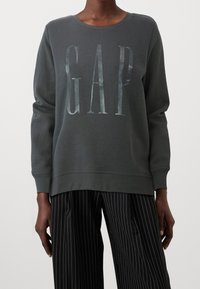 Felpa grigia scura con scollatura a girocollo e il logo "GAP" in lettere metalliche. Caratterizzata da maniche lunghe e polsini a costine. Indossata con pantaloni a righe.