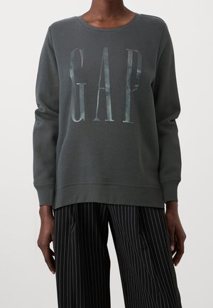 Felpa grigia scura con scollatura a girocollo e il logo "GAP" in lettere metalliche. Caratterizzata da maniche lunghe e polsini a costine. Indossata con pantaloni a righe.