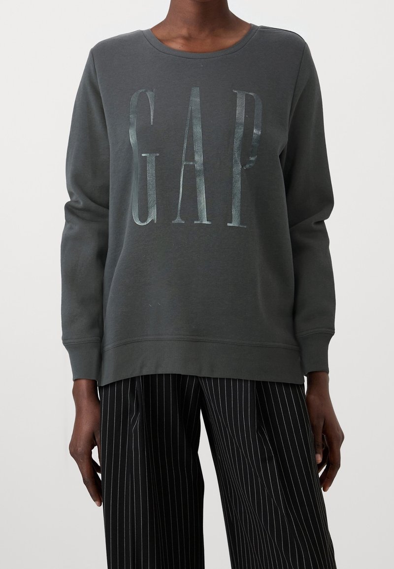 Felpa grigia scura con scollatura a girocollo e il logo "GAP" in lettere metalliche. Caratterizzata da maniche lunghe e polsini a costine. Indossata con pantaloni a righe.