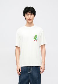 Bijela pamučna majica opuštenog kroja, s šarenim prikazom ukrašene božićne jelke sprijeda. Traper šorc s prugama.