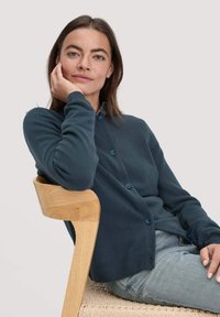 Dunkelblauer Cardigan mit Knopfverschluss, gerippten Manschetten und weicher Textur, getragen über einem Hemd mit Kragen. Modell sitzt auf einem Holzstuhl.