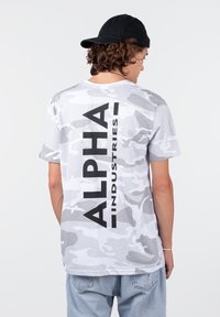 Maskáčové tričko v šedých odstínech. Na zádech se nachází výrazný černý nápis "ALPHA INDUSTRIES" ve vertikální poloze. Krátké rukávy, uvolněný střih, bavlněný materiál.