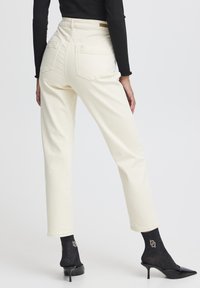 Jeans de color crema, de cintura alta, con pierna recta, bolsillos traseros y una textura suave, combinados con unos zapatos de tacón negros, brillantes y de punta, y calcetines.