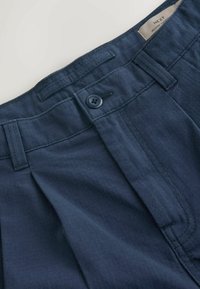 Navy blue trousers with a textured fabric, featuring a button closure and front pleats. Label visible at the waistband's interior.