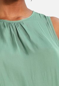 Zelená bezrukávová blůza vyrobená z hladké látky, se zaobleným výstřihem a širokými plisovanými detaily v přední části, což vytváří volný a splývavý střih.