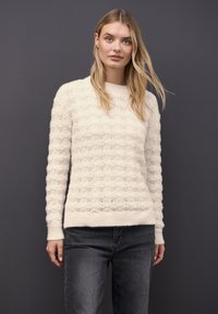 Suéter de punto color crema con un patrón de ondas texturizado, cuello redondo y dobladillo acanalado. Combinado con jeans de mezclilla grises.