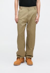 Pantaloni largi khaki dintr-un material robust, cu buzunare frontale, o textură netedă și o croială lejeră, asortați cu cizme de lucru maro.