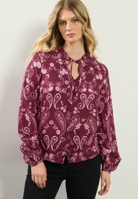 Blusa bordeaux con motivo floreale e paisley, con scollatura a chiave, maniche lunghe con polsini arricciati e chiusura con bottoni.