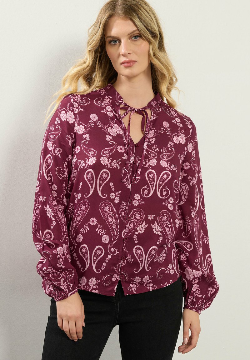 Blusa bordeaux con motivo floreale e paisley, con scollatura a chiave, maniche lunghe con polsini arricciati e chiusura con bottoni.